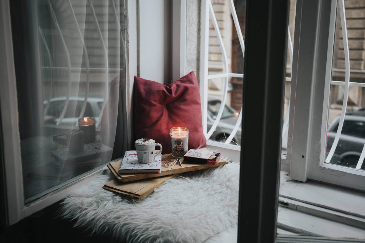 A red cushion, scented candle, a box of matches, a cup of coffee on a book, over two wooden planks placed over each other, all on a small fluffy mat near the window