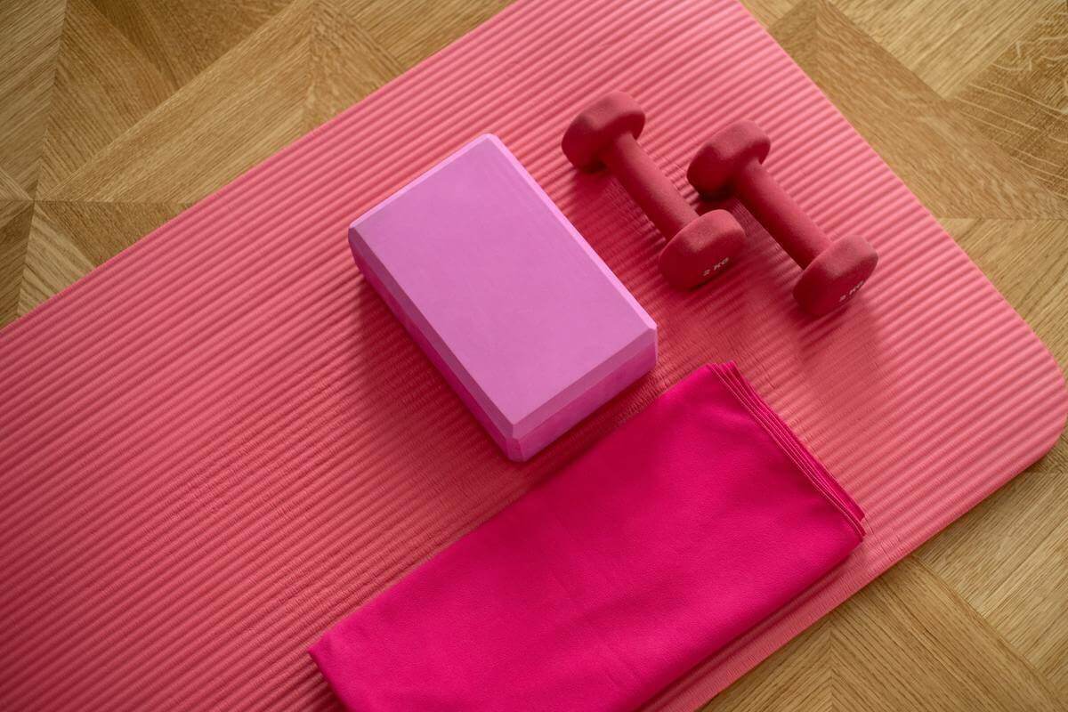 A pair of pink dumbbells, a pink box and a pink towel on a pink yoga mat laid on wood flooring